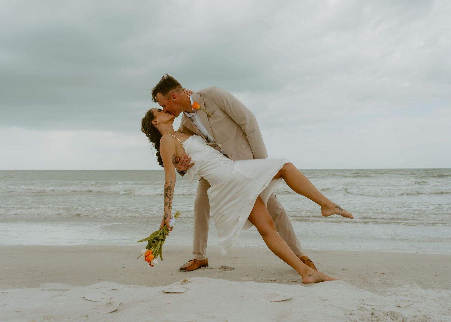Couple sharing romantic dip kiss on sandy beach during wedding, bride holding colorful bouquet with ocean backdrop.