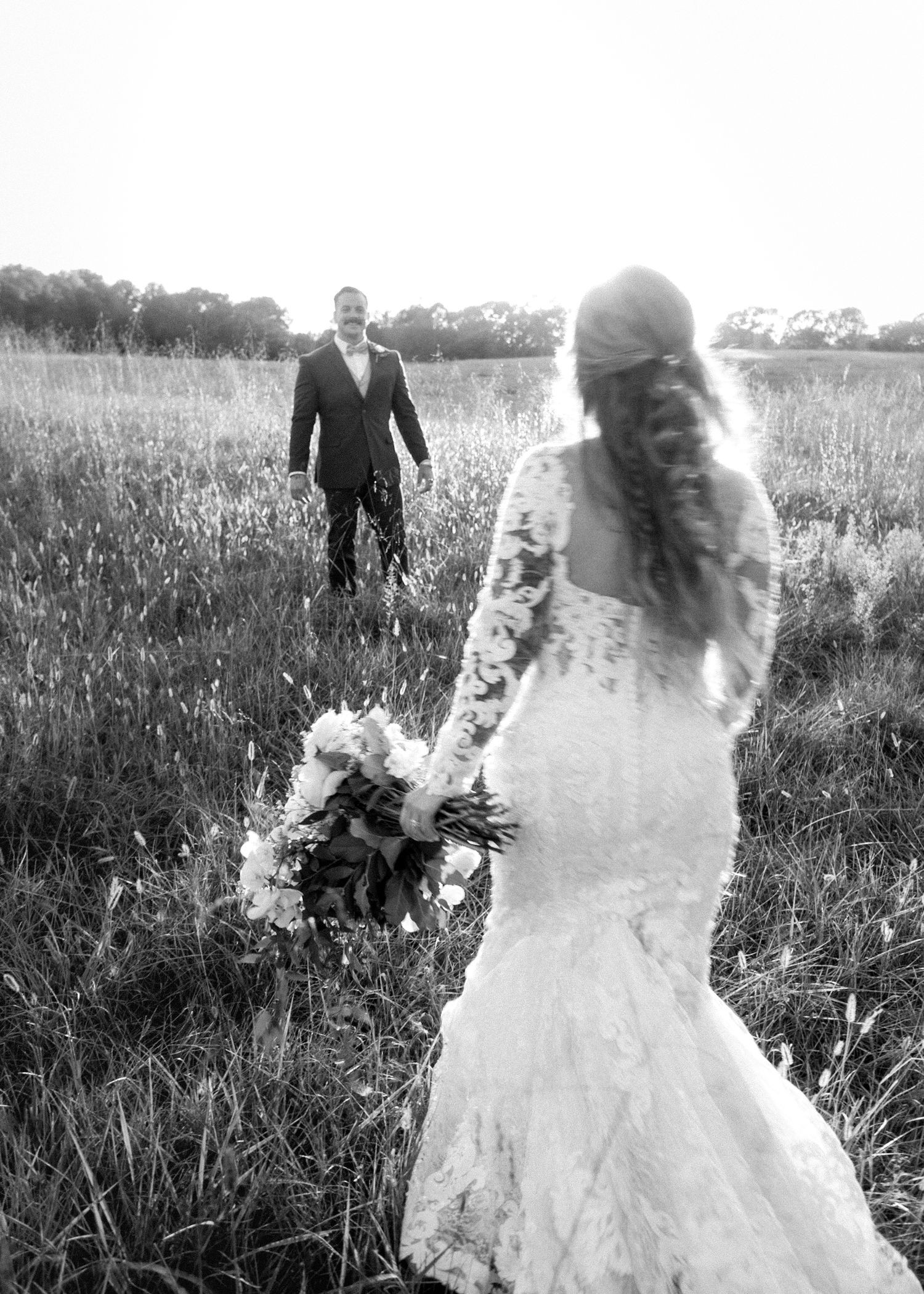 Black and white photo of a bride in lace gown holding bouquet as groom walks toward her in a field.