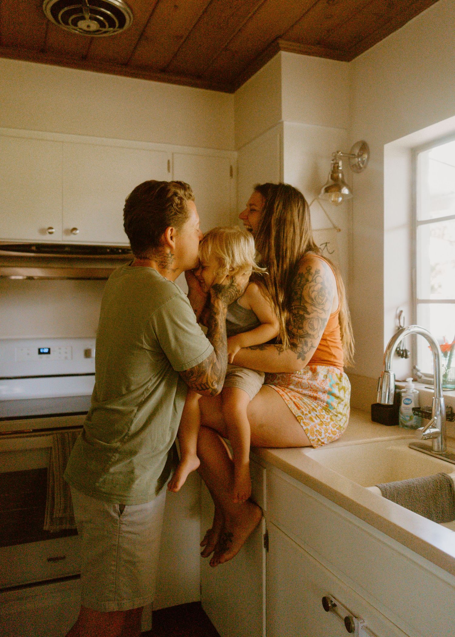 A tattooed couple shares a tender moment with their toddler in a cozy kitchen, the child sitting on the counter.