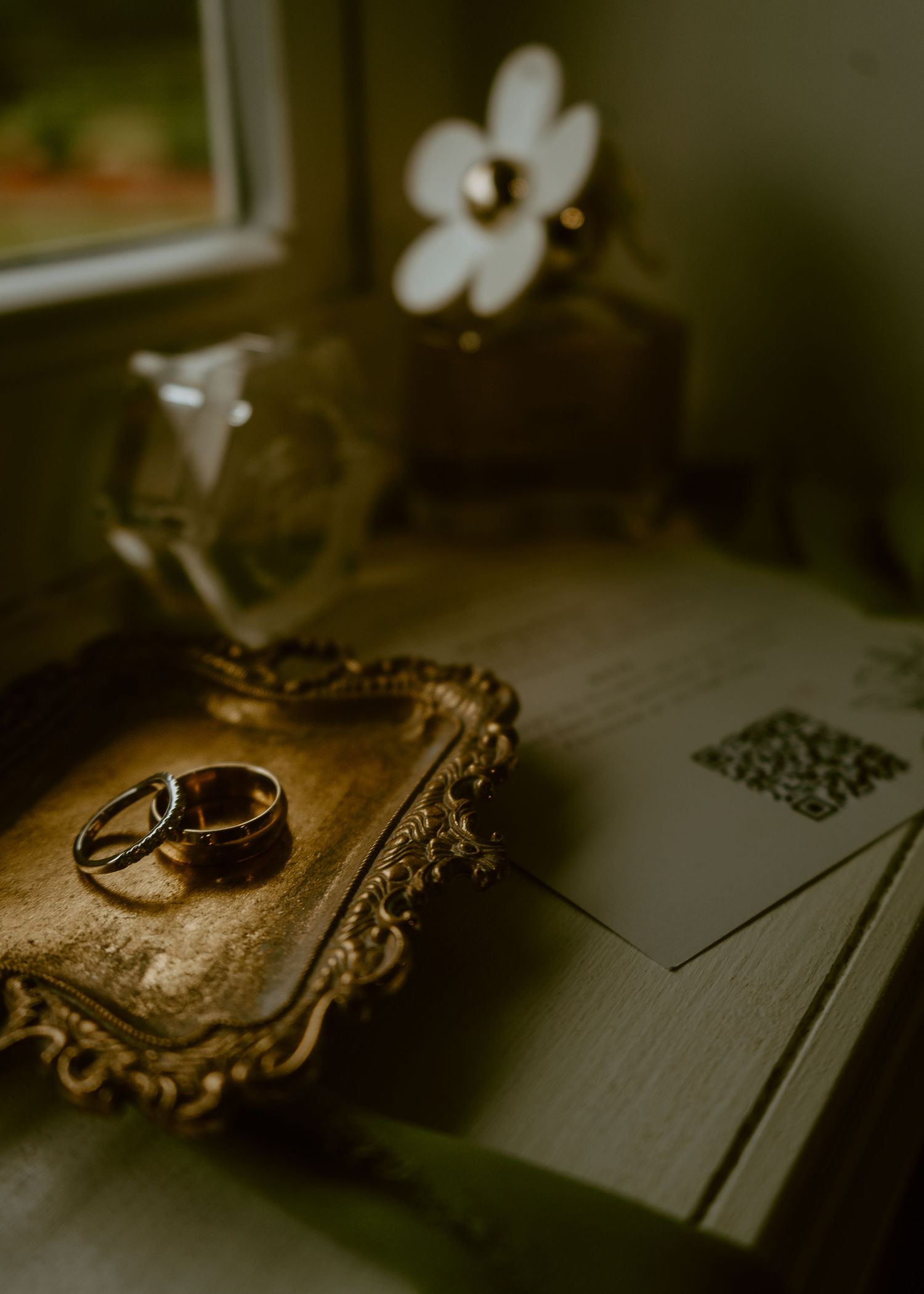 Two gold wedding bands resting on an ornate vintage tray beside wedding invitations and a QR code card near a window.