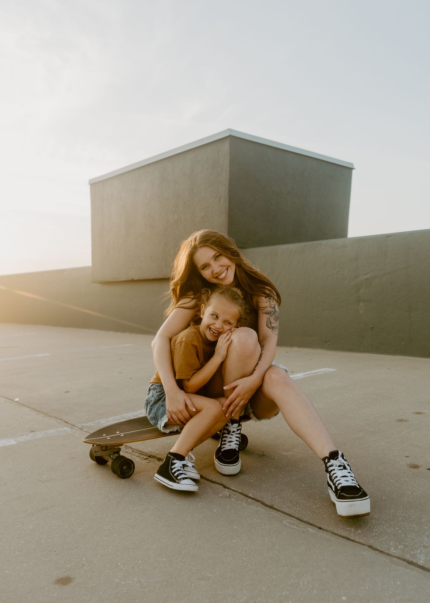 Woman and child sitting on a skateboard on a rooftop at golden hour, smiling and embracing warmly.