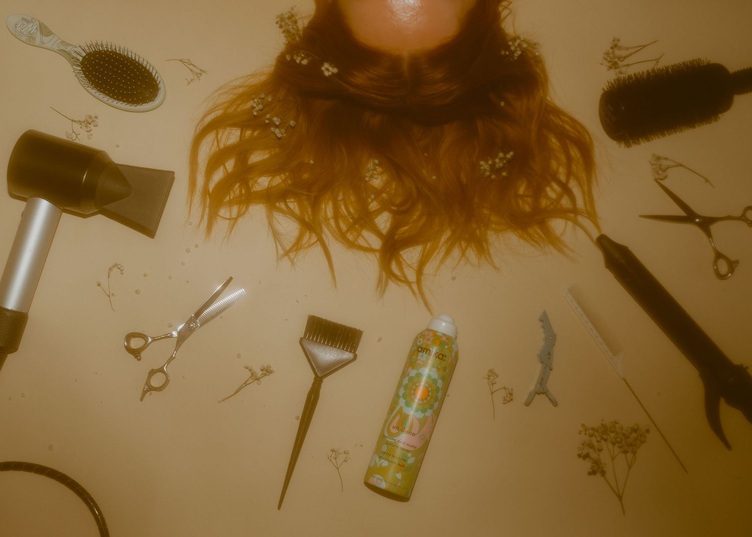 Overhead view of red hair surrounded by salon tools including scissors, hair dryer, dye brush, and spray on beige surface.