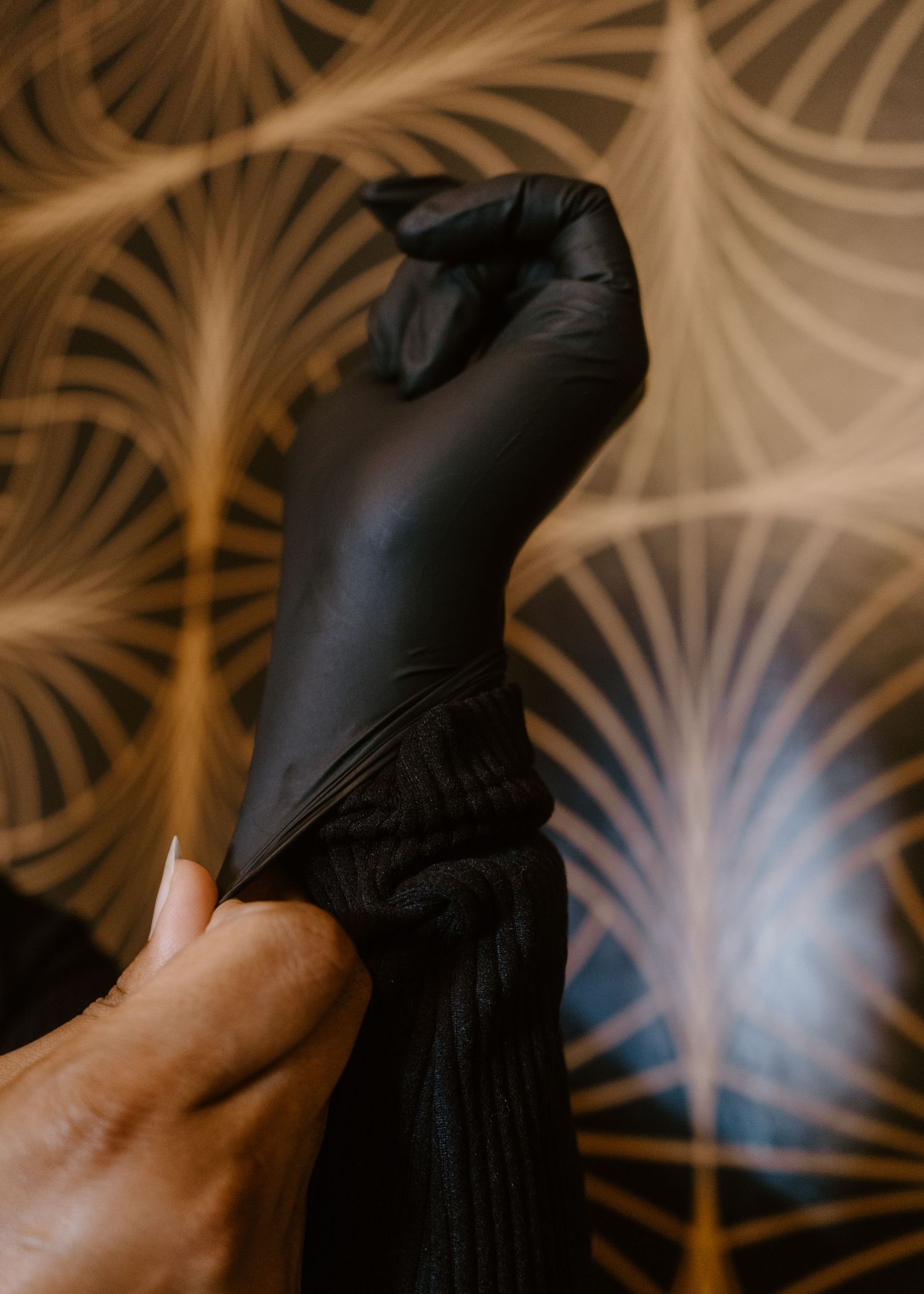 Hands pulling on a black nitrile glove against a decorative gold leaf patterned wallpaper background.