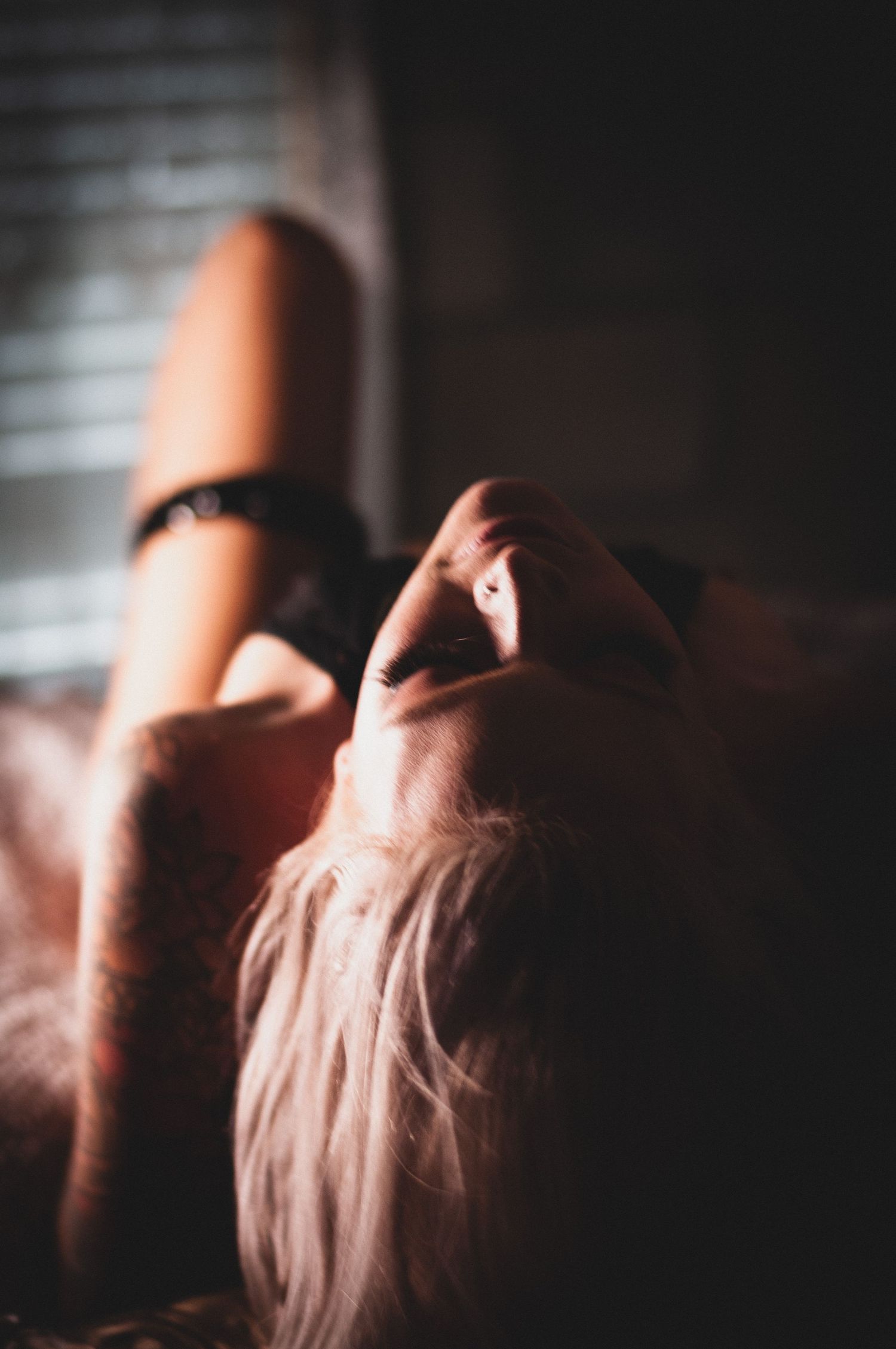 A woman with long blonde hair reclines in moody, dramatic lighting near window blinds wearing black lingerie.
