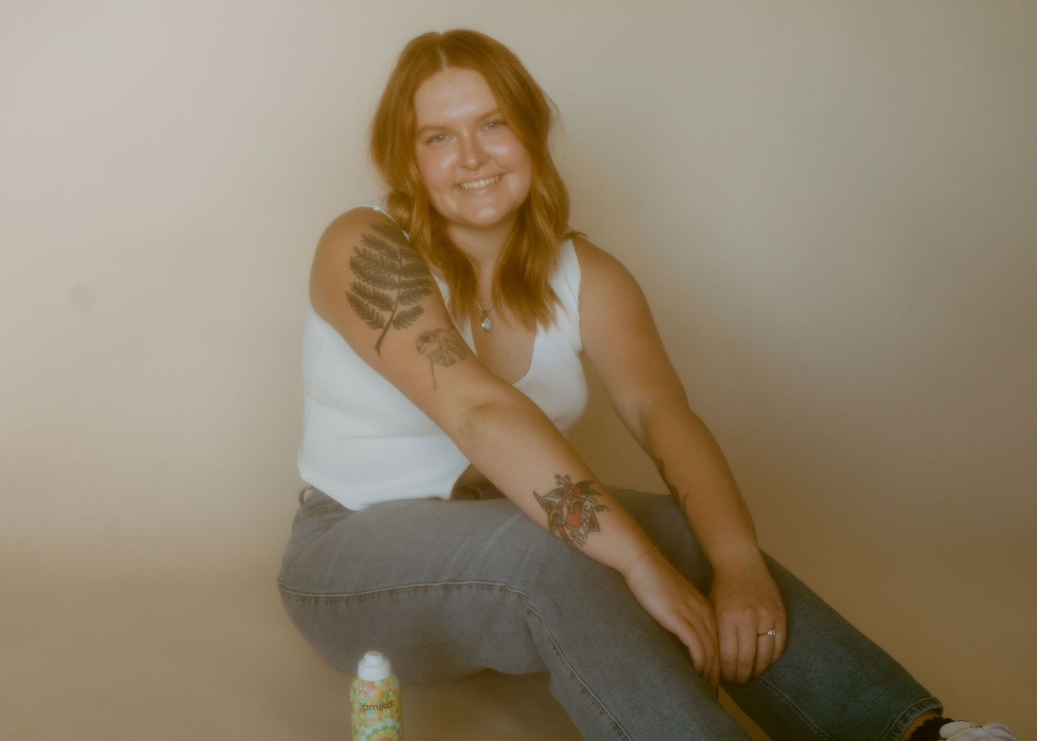 Young woman with tattoos sitting casually in white tank top and jeans, smiling against a neutral background.