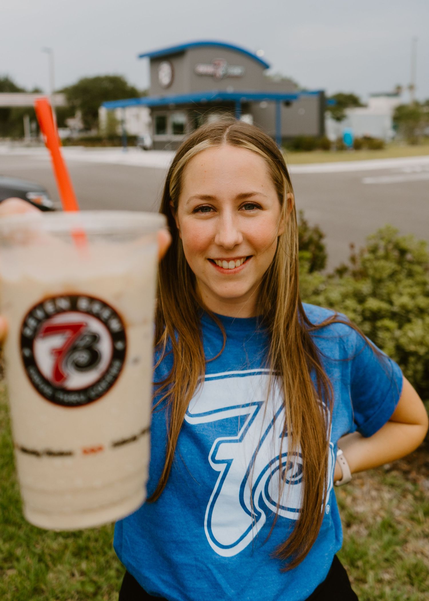 Smiling woman holding a large iced drink with red straw, wearing a blue 7 Brew Coffee shirt outdoors.