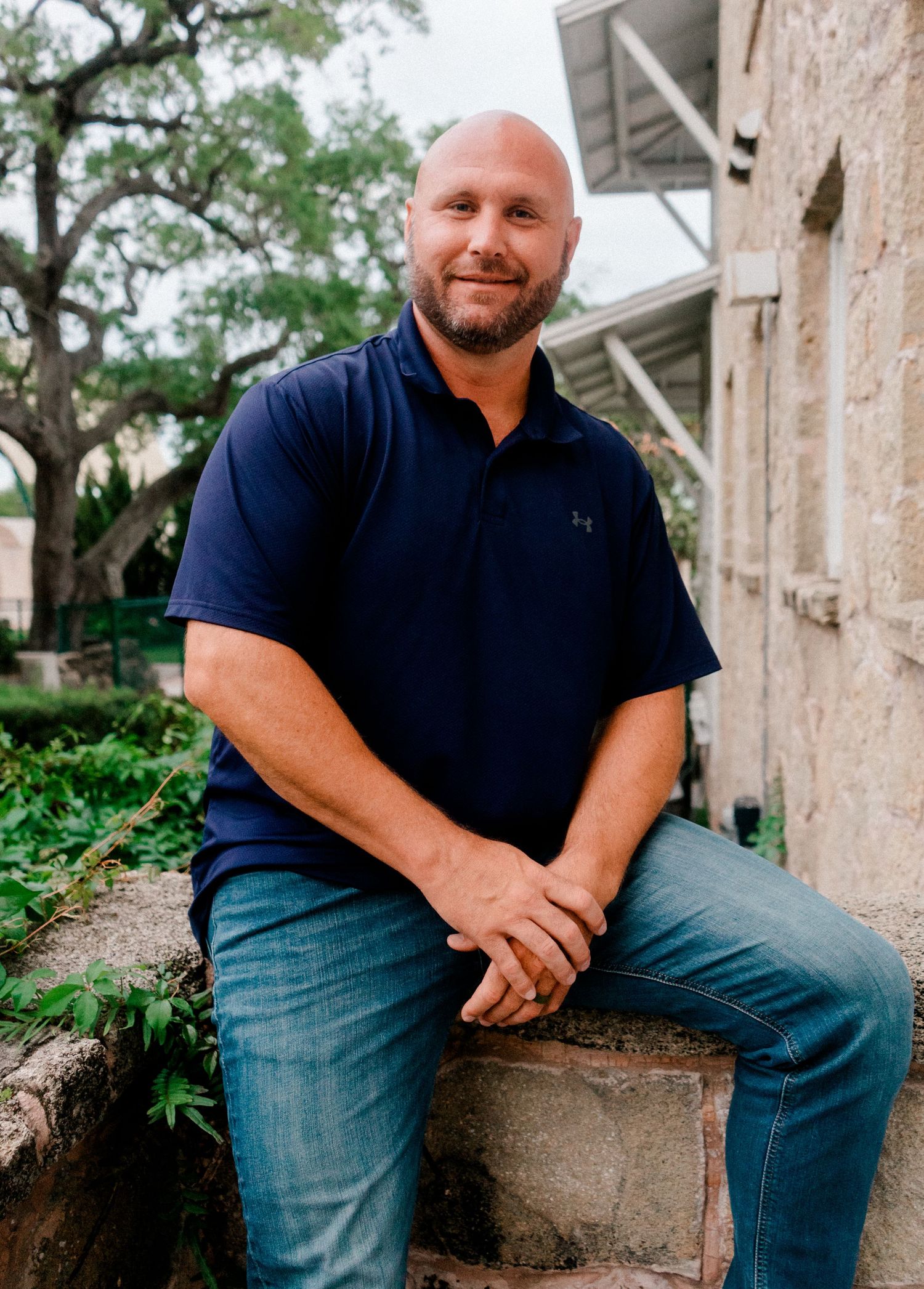 Smiling bald man with beard wearing navy polo shirt sitting on stone wall outdoors near historic building.