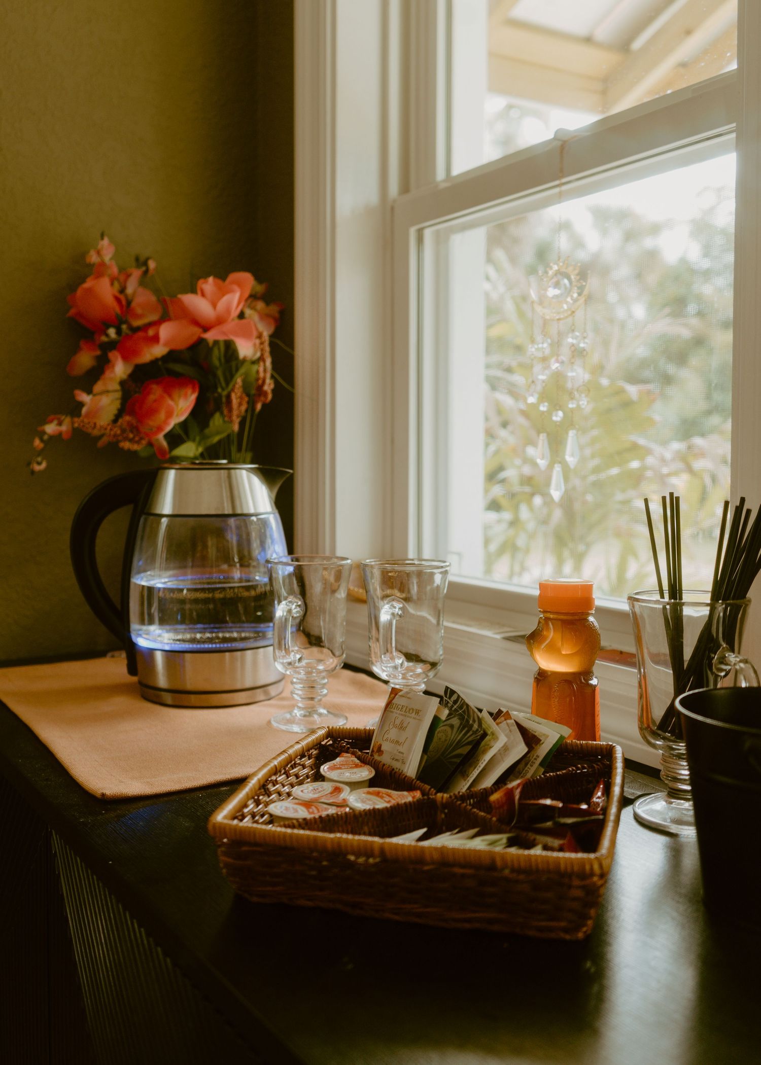 Cozy window nook with electric kettle, pink flowers, wine glasses, wicker tray of teas, and candle on wooden table.