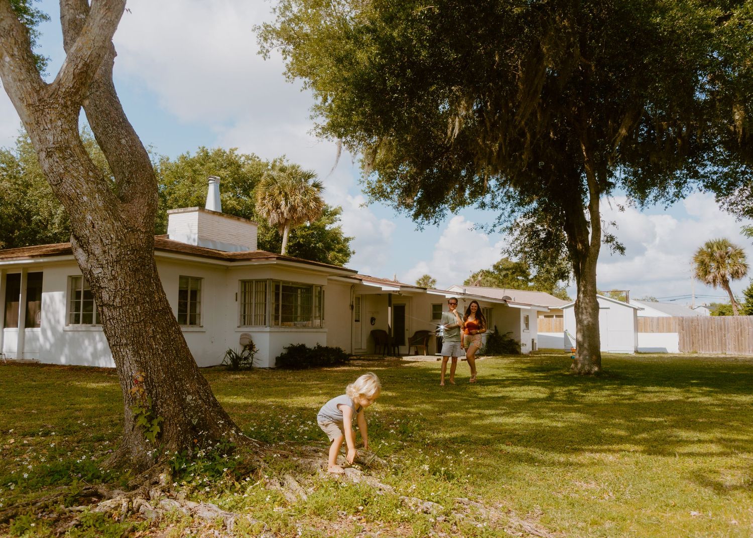 Children playing in a sunny yard with large trees in front of a white single-story Florida home.