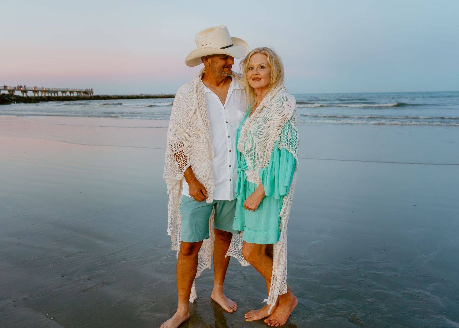 Elderly couple in matching white and turquoise outfits posing on a beach at sunset.