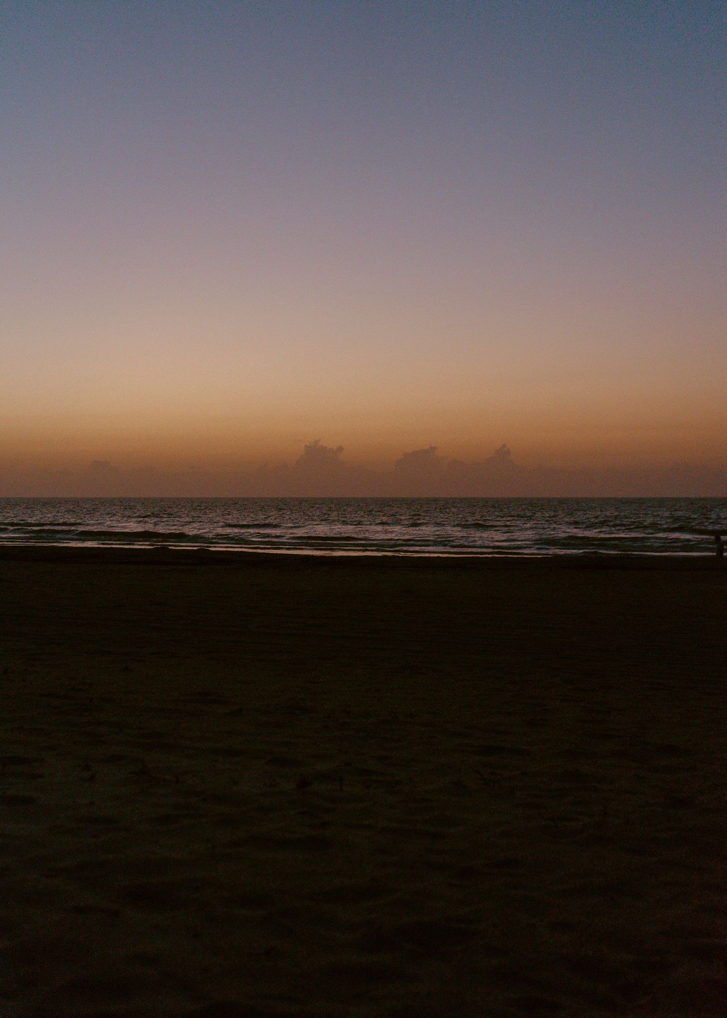 Serene beach sunset with warm orange and pink hues over calm ocean waves and dark sandy shore.