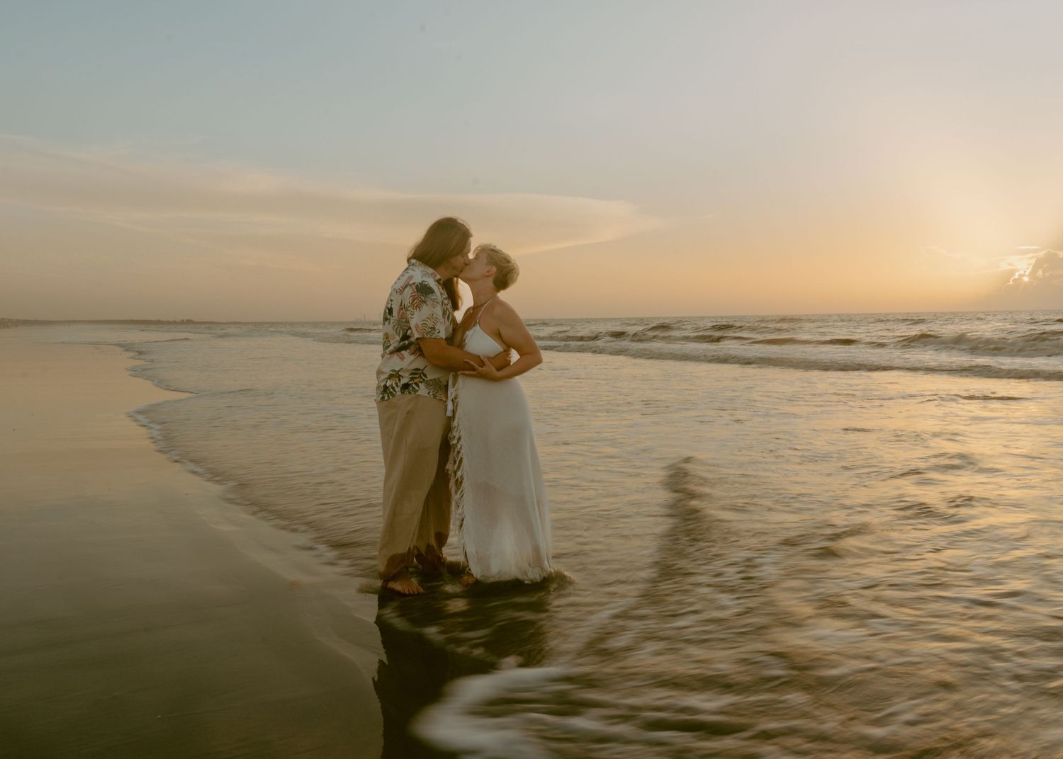 A couple shares a romantic kiss on a sandy beach at golden sunset, waves gently lapping around their feet.
