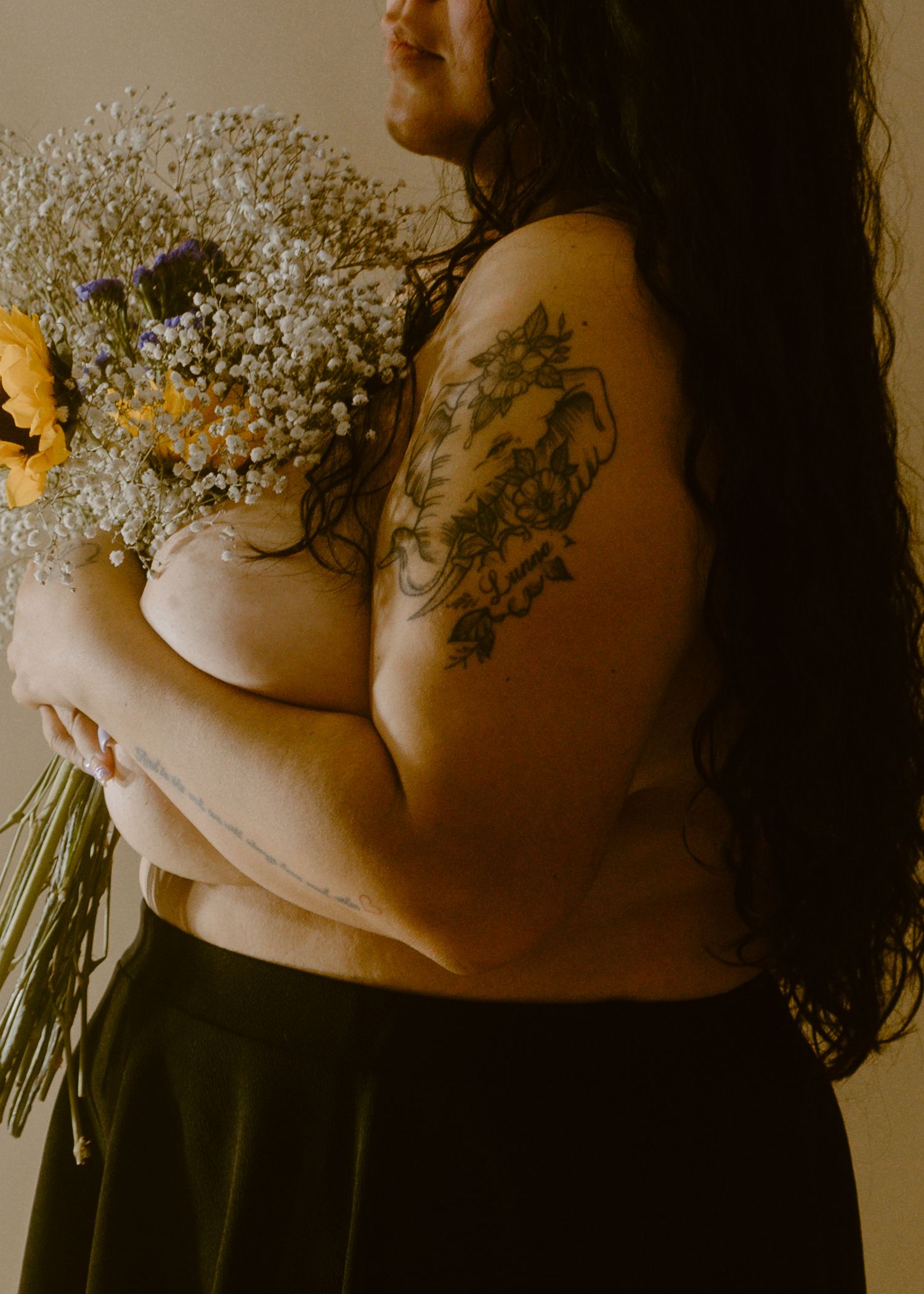 Plus-size woman with floral tattoo holding yellow wildflower bouquet, artistic boudoir photography.
