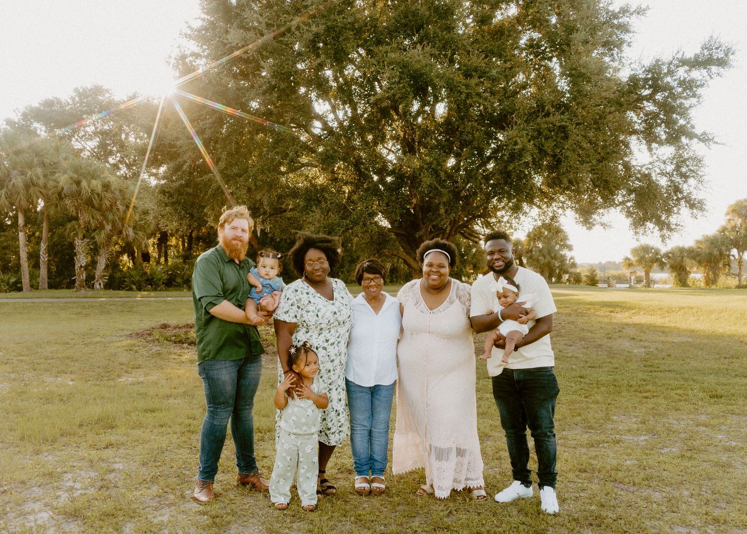 Multi-generational family portrait outdoors under a large oak tree during golden hour sunset in a green park.