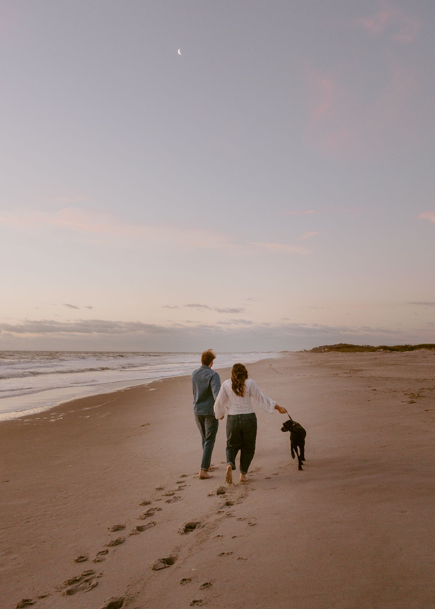 Couple walking barefoot on a sandy beach at sunset with their black dog, footprints in sand, crescent moon visible.