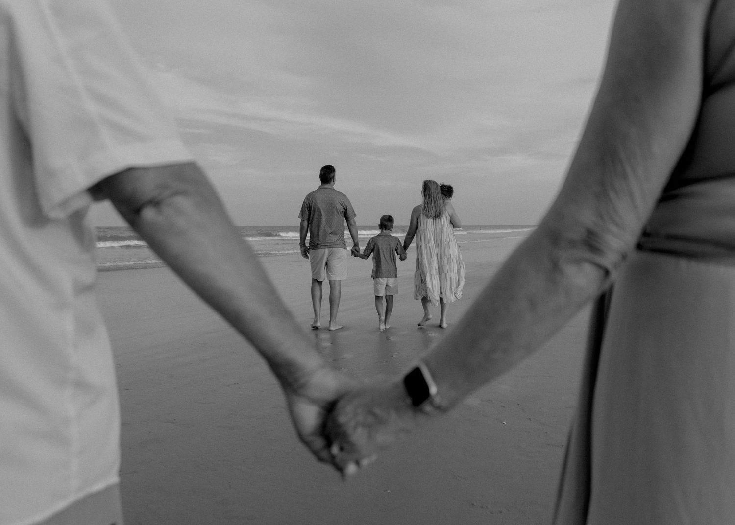 Black and white photo of a family holding hands on a beach, walking toward the ocean waves together.