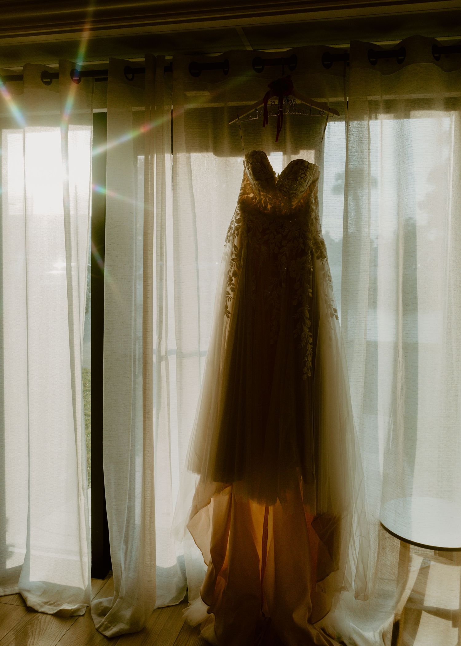 A beaded strapless wedding gown hangs elegantly in a sunlit window with sheer white curtains and a warm lens flare.