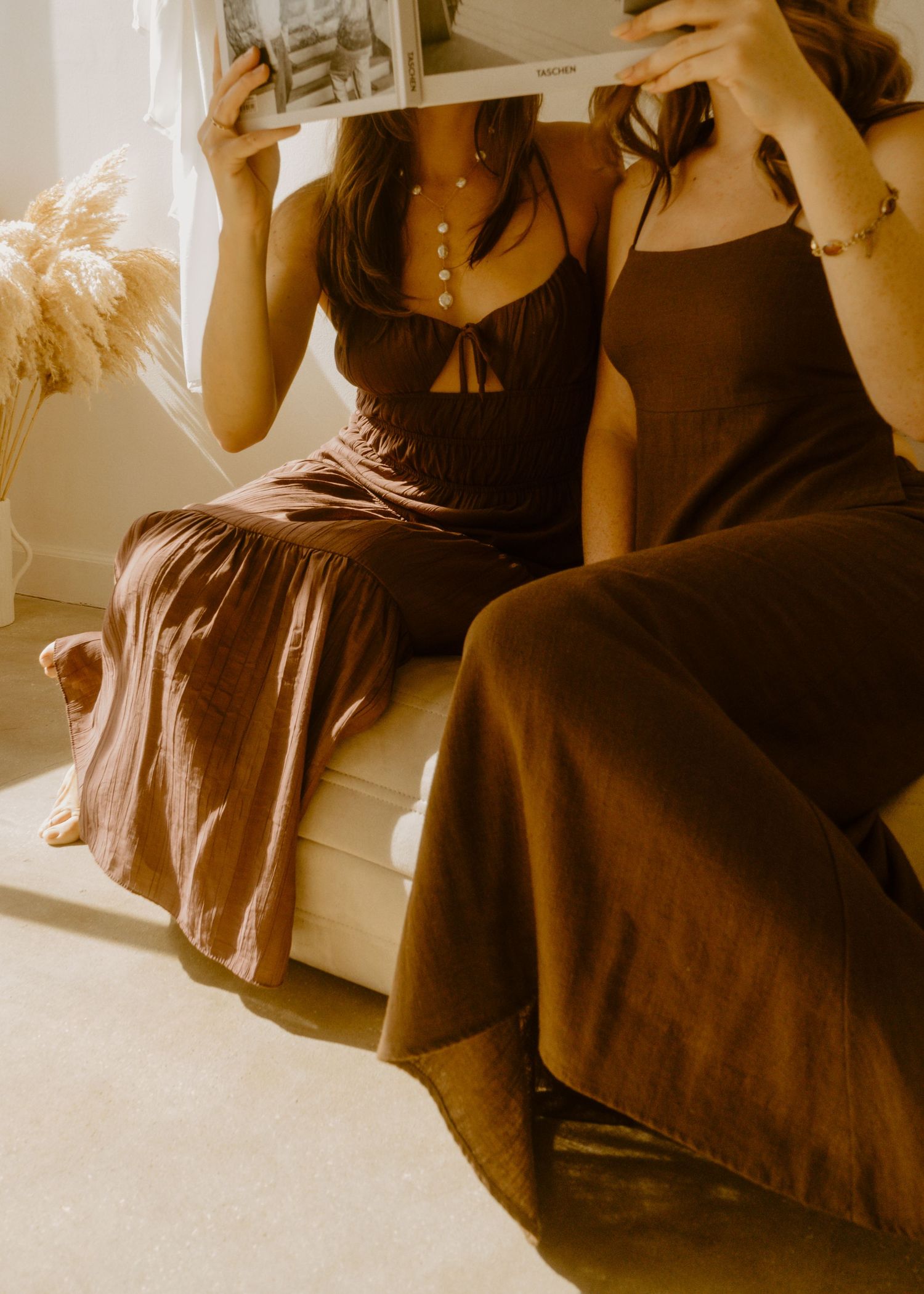 Two women in brown dresses sit together, faces hidden behind a magazine, in a warm, sunlit room.
