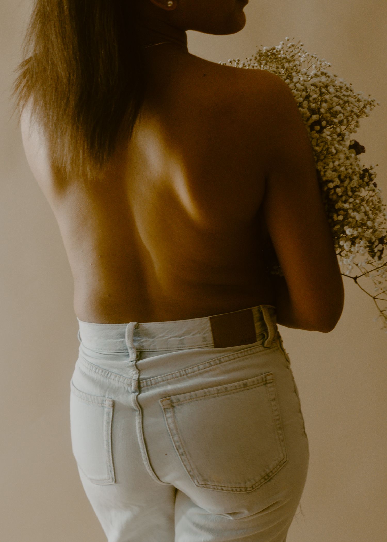 Woman with bare back holding white wildflowers, wearing light wash jeans, photographed from behind.