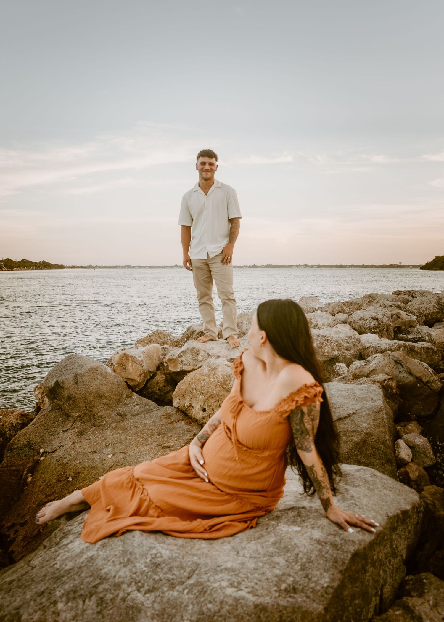 Pregnant woman in orange dress sits on rocks by water as man in white shirt stands behind her at sunset.