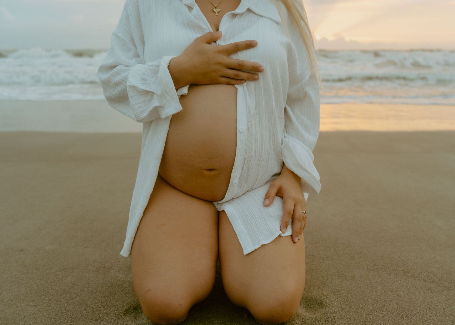 Pregnant woman kneeling on beach sand in open white shirt holding baby bump at golden hour.