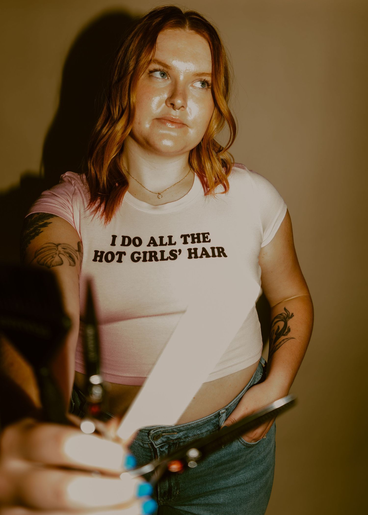 Woman hairstylist in white tee reading I Do All The Hot Girls Hair, holding scissors, with tattoos.