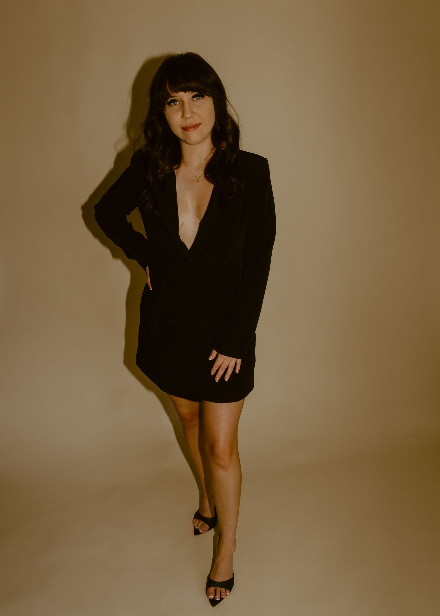 Woman in black blazer mini dress and heels posing confidently against a warm beige background.