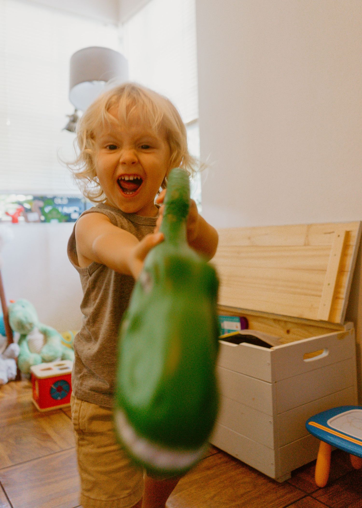 Excited toddler with curly blonde hair pointing a green toy dinosaur at the camera in a playroom.