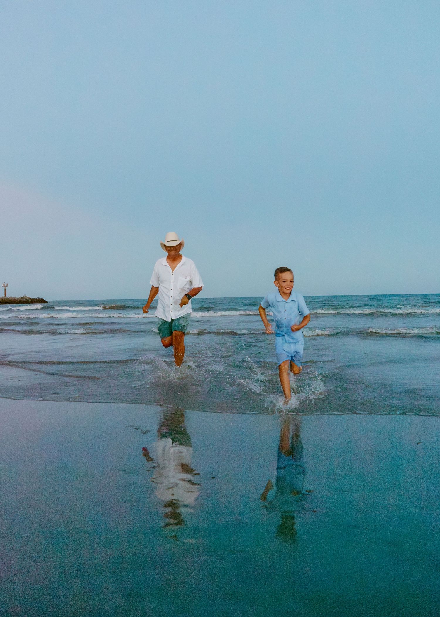 A man and young boy running joyfully through shallow ocean waves on a sunny beach.