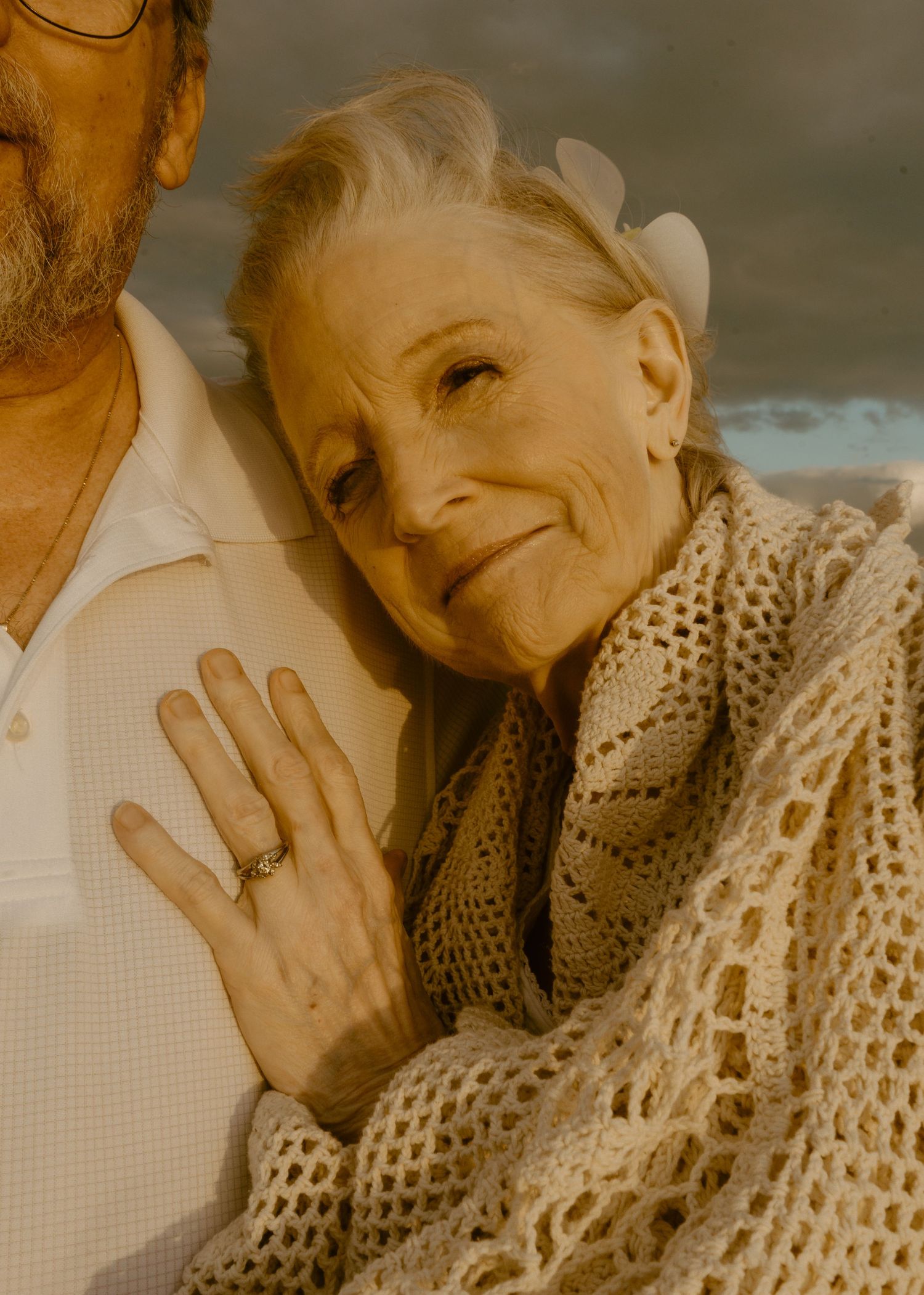 Elderly woman smiling warmly wrapped in knit shawl with flower in hair leaning on partner.