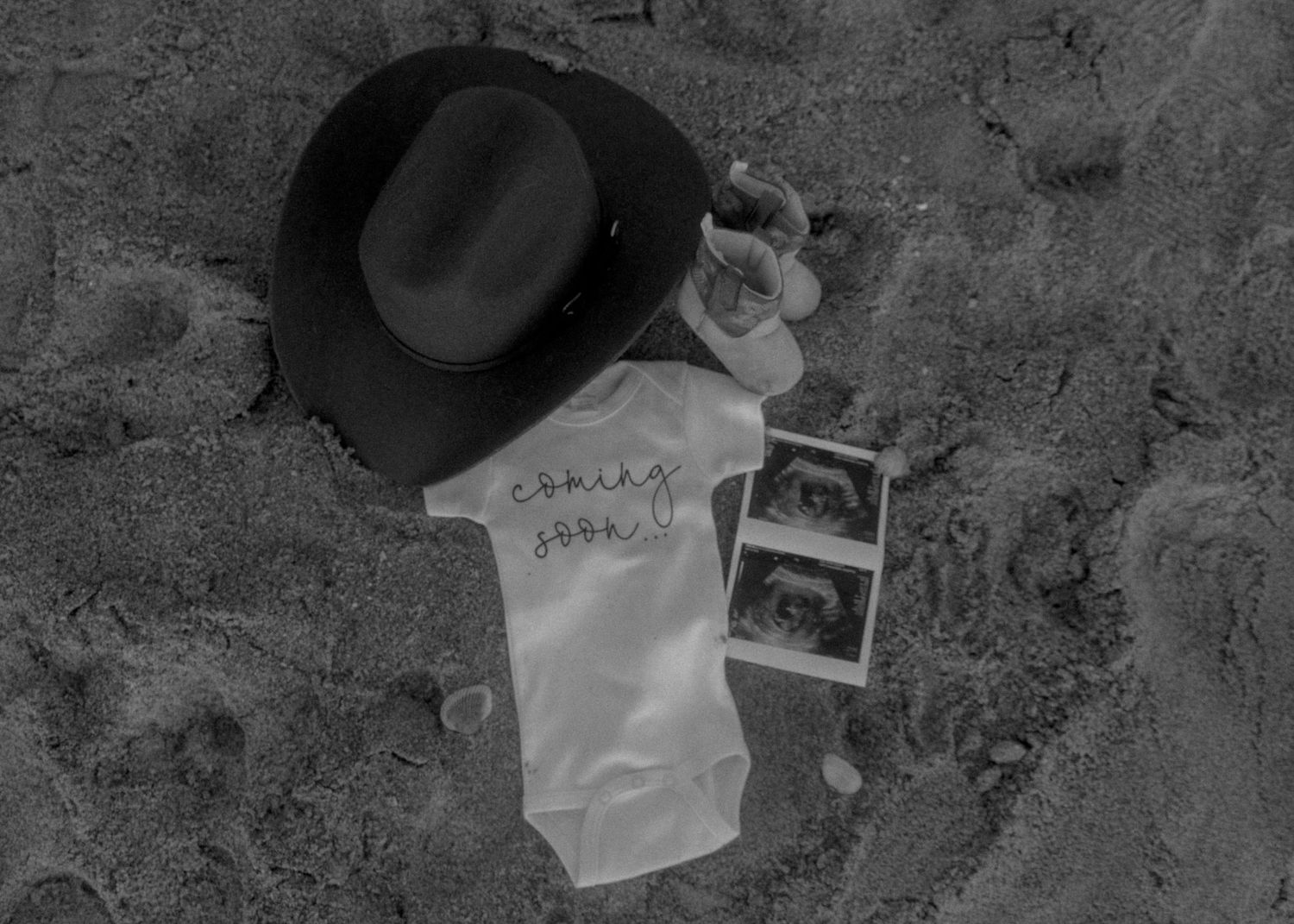 Black and white flat lay of baby onesie, hat, shoes, and ultrasound on sand.
