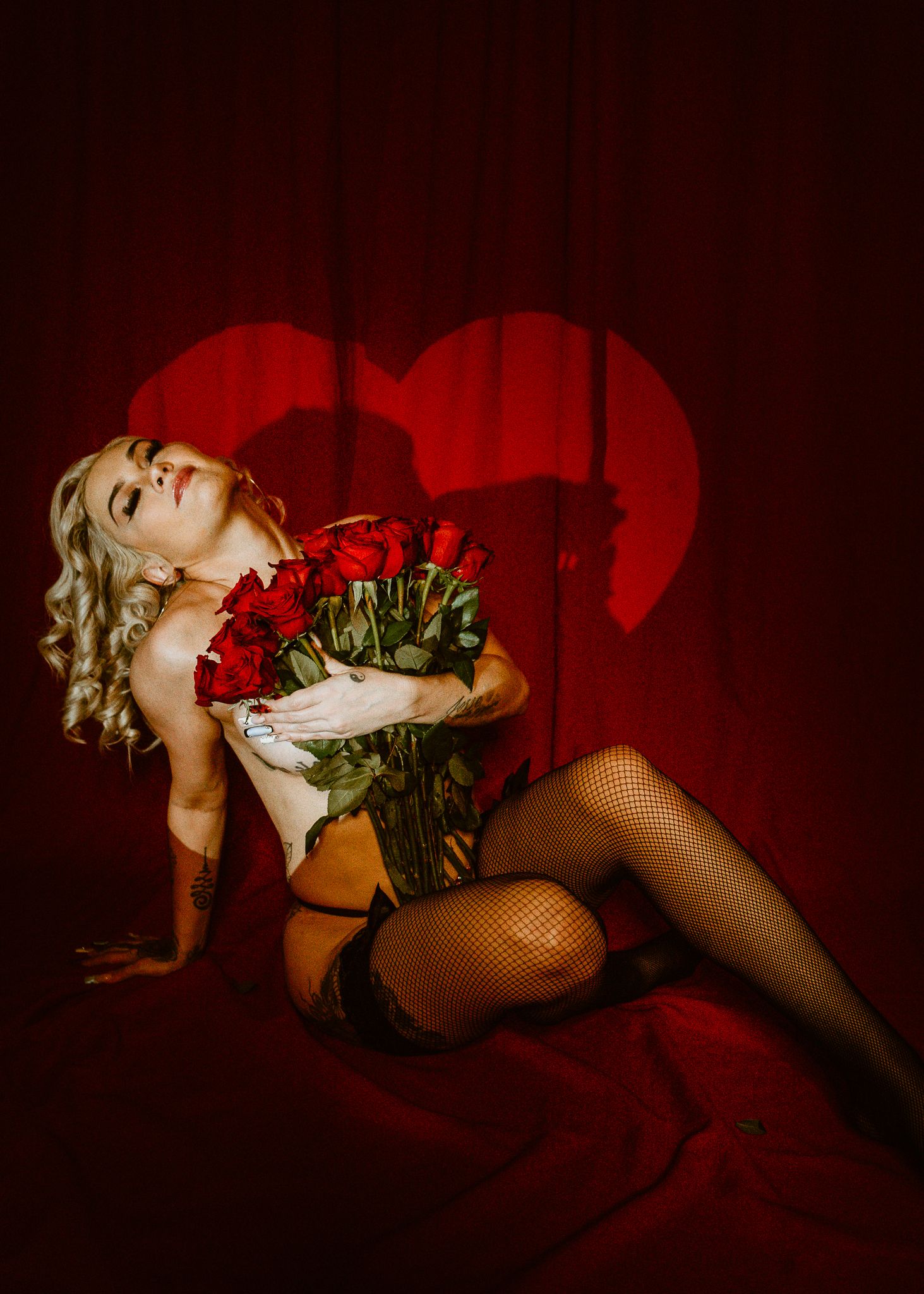 Blonde woman in lingerie and fishnet stockings poses with red roses against a dramatic red heart shadow backdrop.