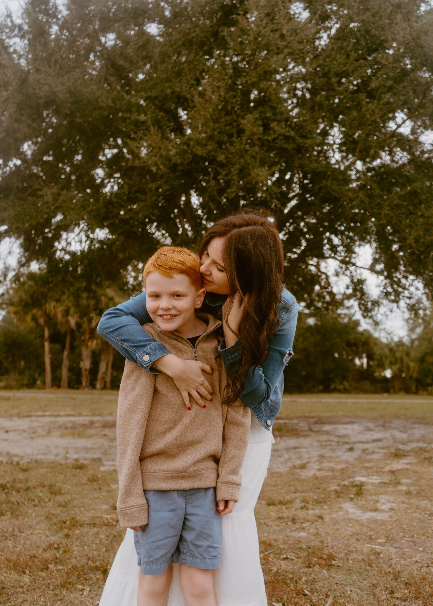 A mother kisses her red-haired son outdoors near trees in a warm, candid family portrait session.