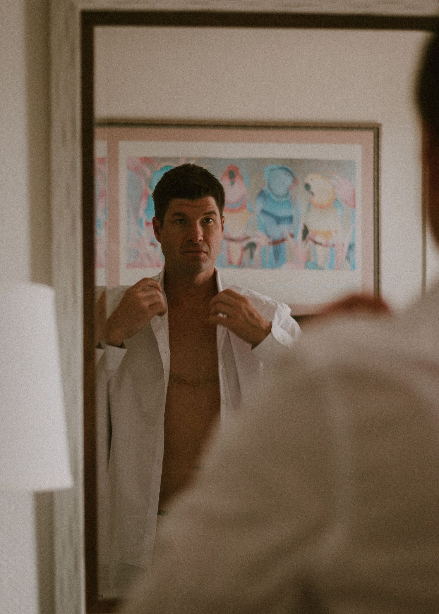 Man buttoning white shirt in mirror reflection with colorful artwork hanging on wall behind him.