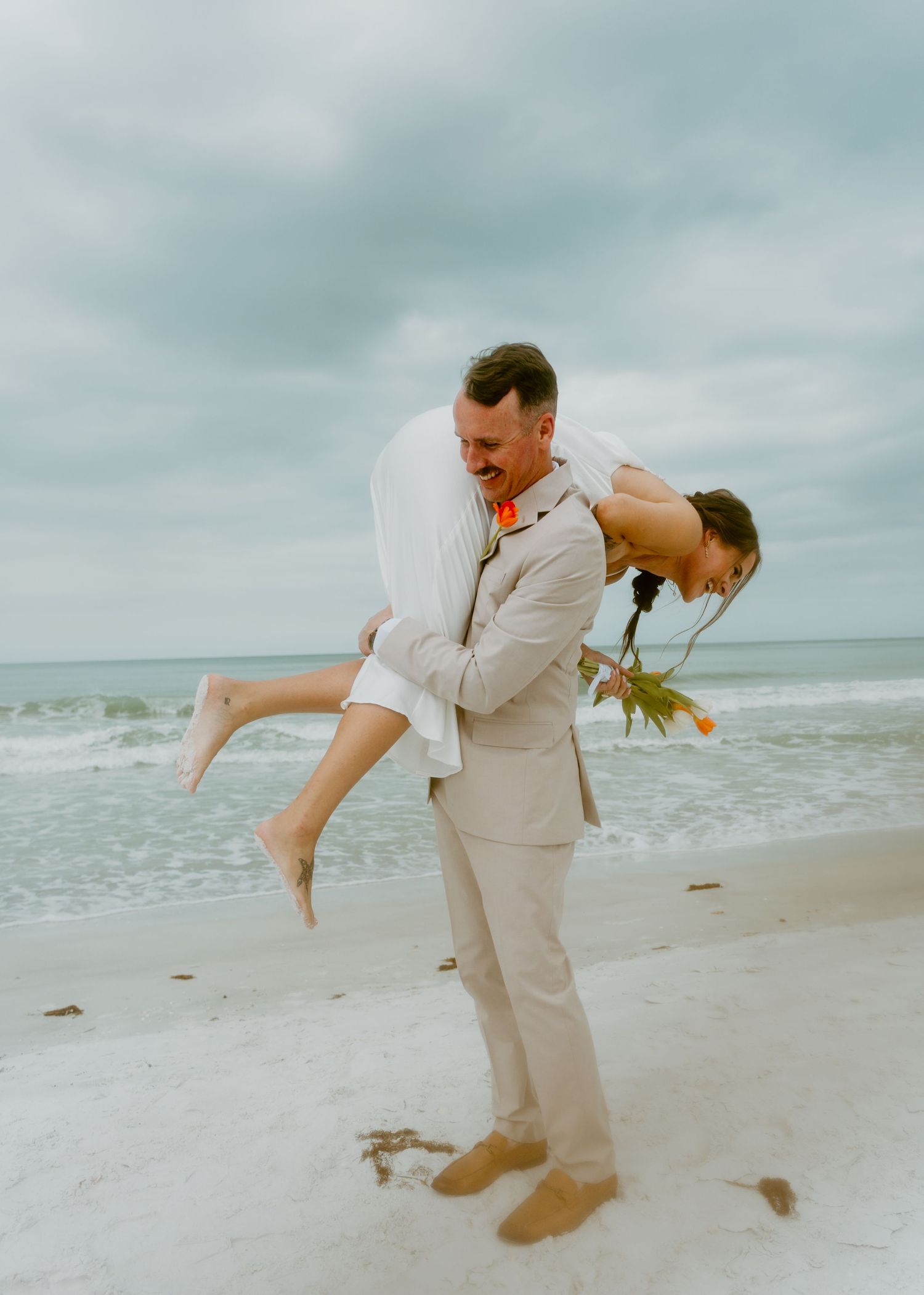 Groom lifting bride on beach wedding, ocean waves background, sandy shore, casual attire, romantic moment.