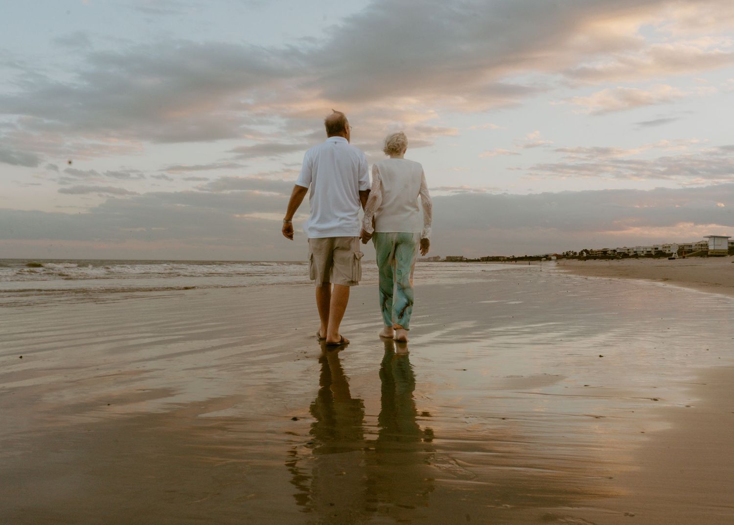Elderly couple walking hand in hand on a reflective beach at sunset, enjoying a peaceful seaside stroll.