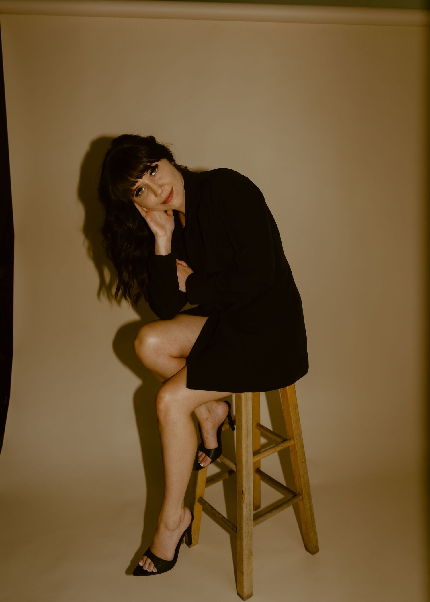 Woman in black blazer dress poses on wooden stool against beige backdrop, wearing heels with dark hair and bangs.