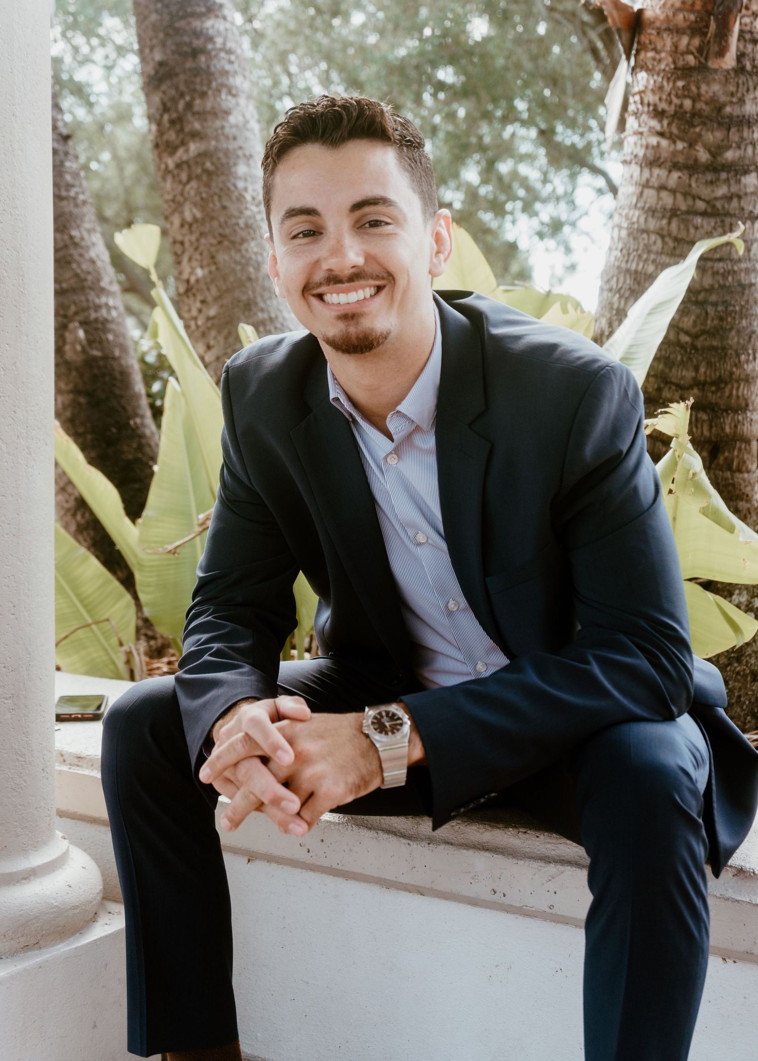 Smiling young man in navy suit sitting outdoors near palm trees for a professional headshot photo.