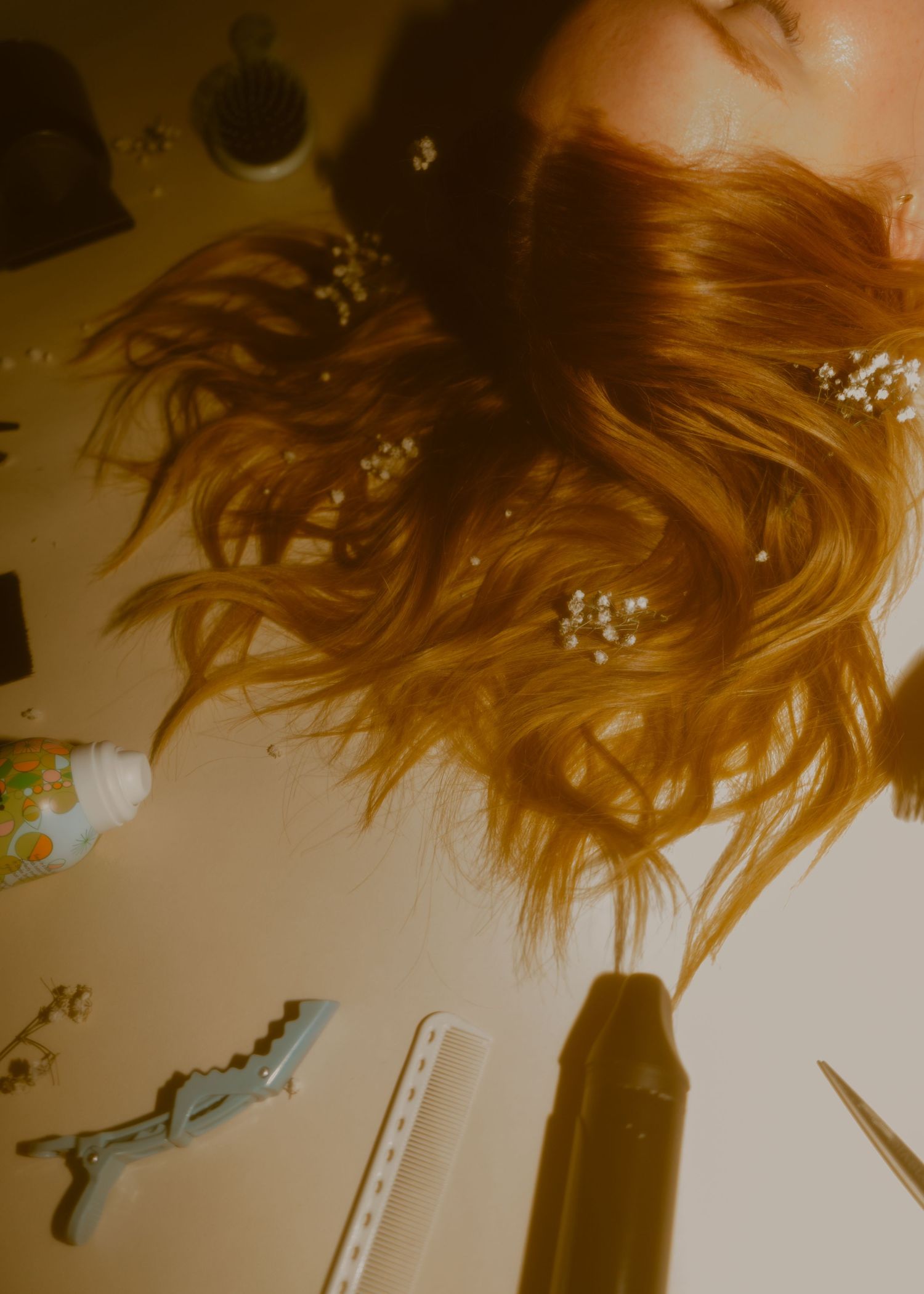 Overhead view of a person with long red hair adorned with small white flowers, surrounded by beauty tools on a table.