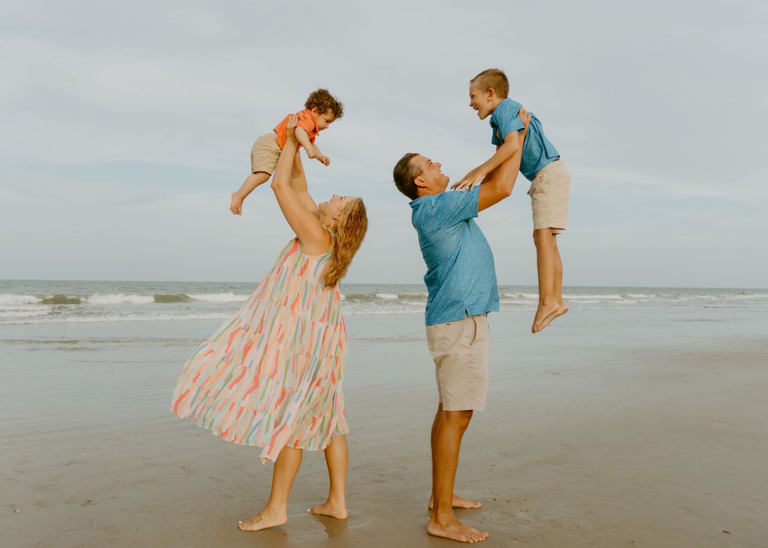 A joyful family plays on the beach, parents lifting two young children in the air near ocean waves.