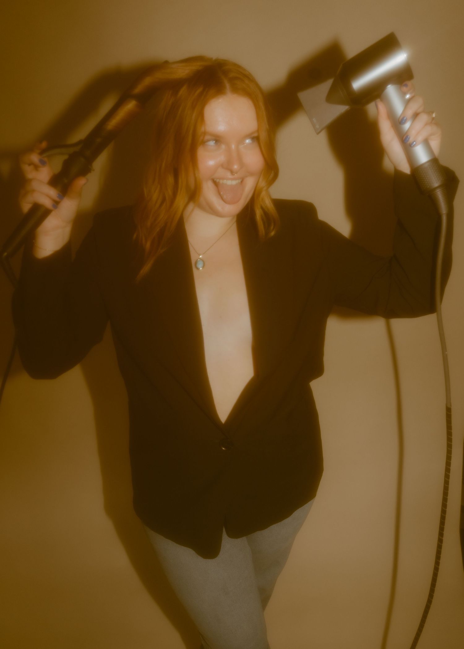 Woman with red hair styling her hair using a curling iron and hair dryer, wearing a black blazer.