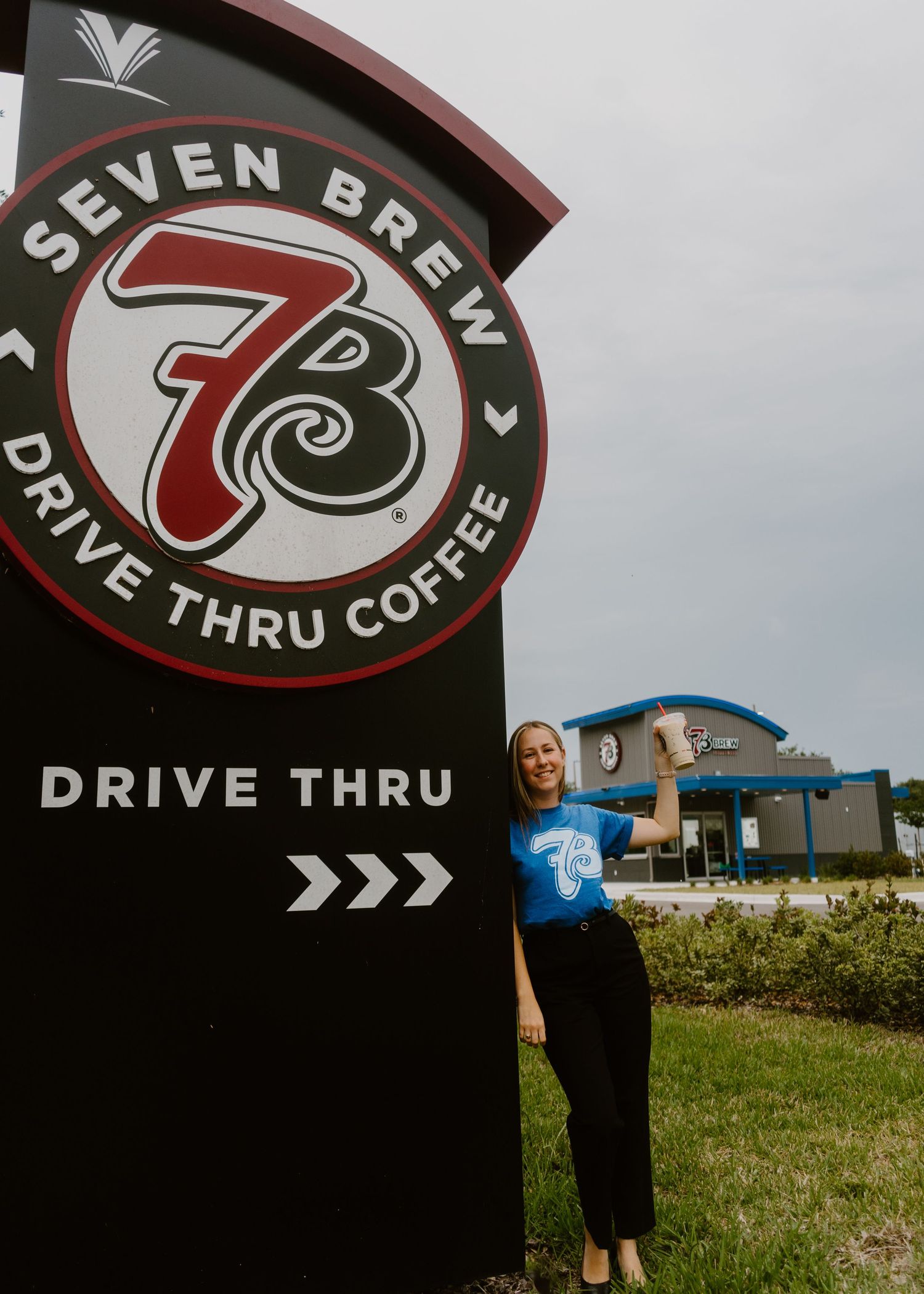 Woman in blue Seven Brew shirt holds coffee cup beside Seven Brew Drive Thru Coffee sign outside location.