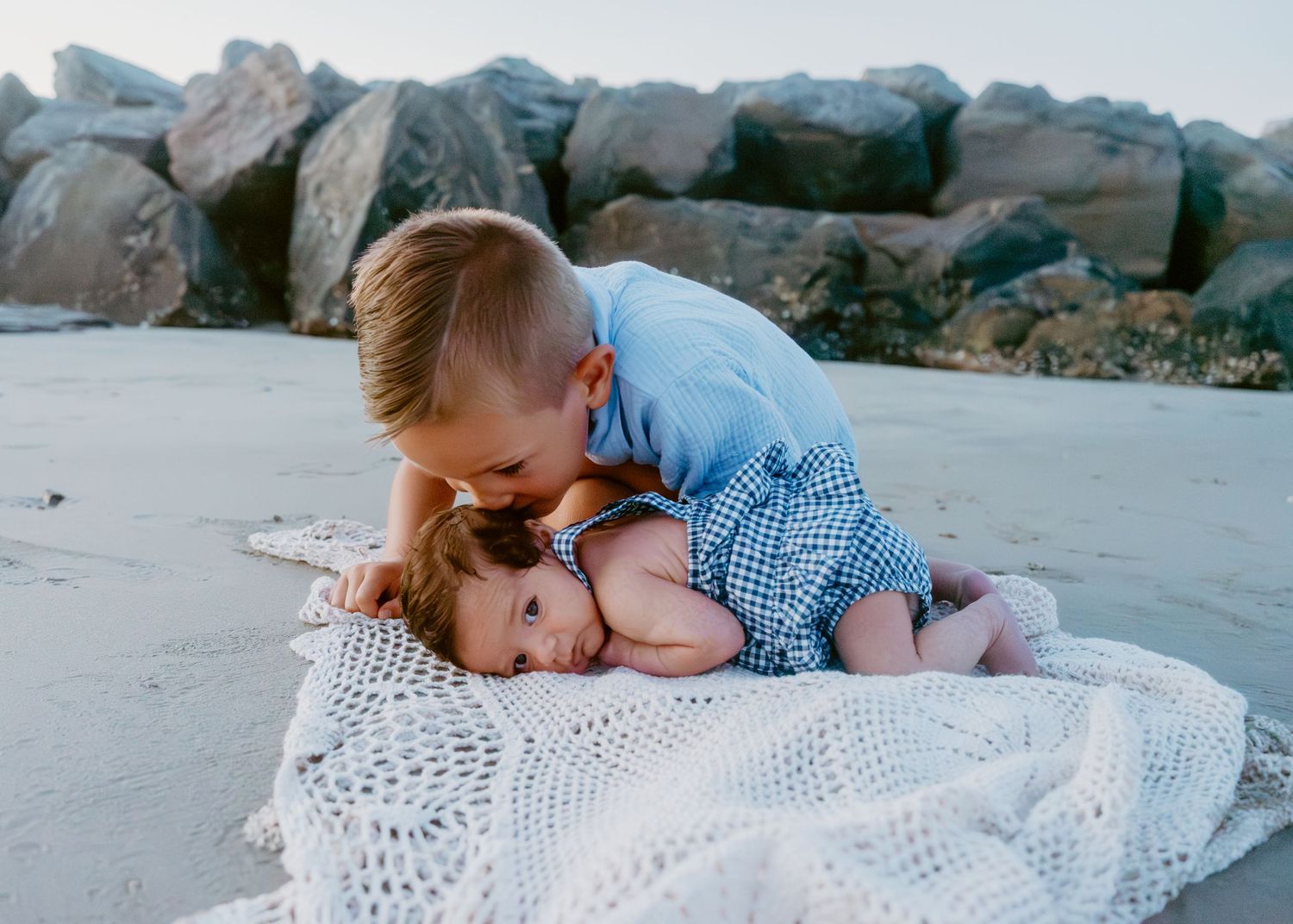 Two young siblings share a sweet moment on a white blanket at a rocky beach, older boy hugging baby girl.