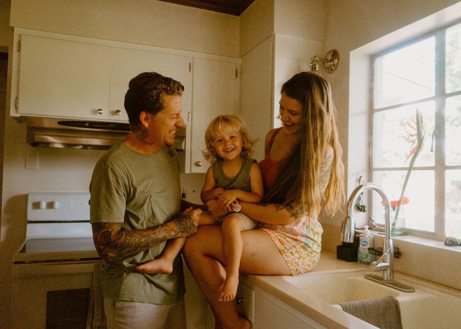 Happy family with tattooed dad, mom, and blonde toddler sitting on kitchen counter by sunny window.
