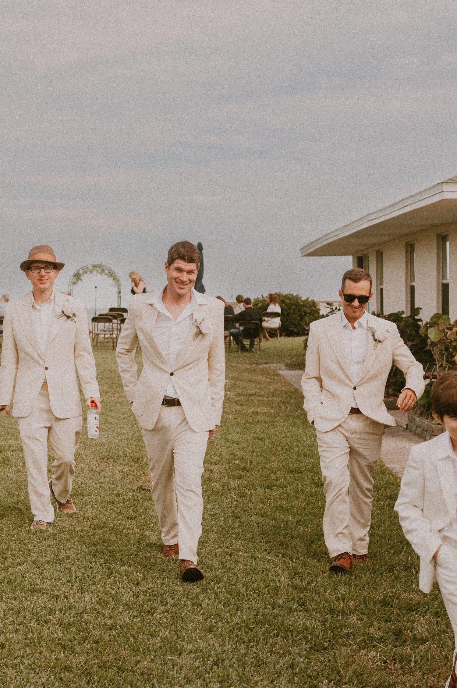 Groomsmen in cream suits walk outdoors at a seaside wedding venue on a cloudy day.