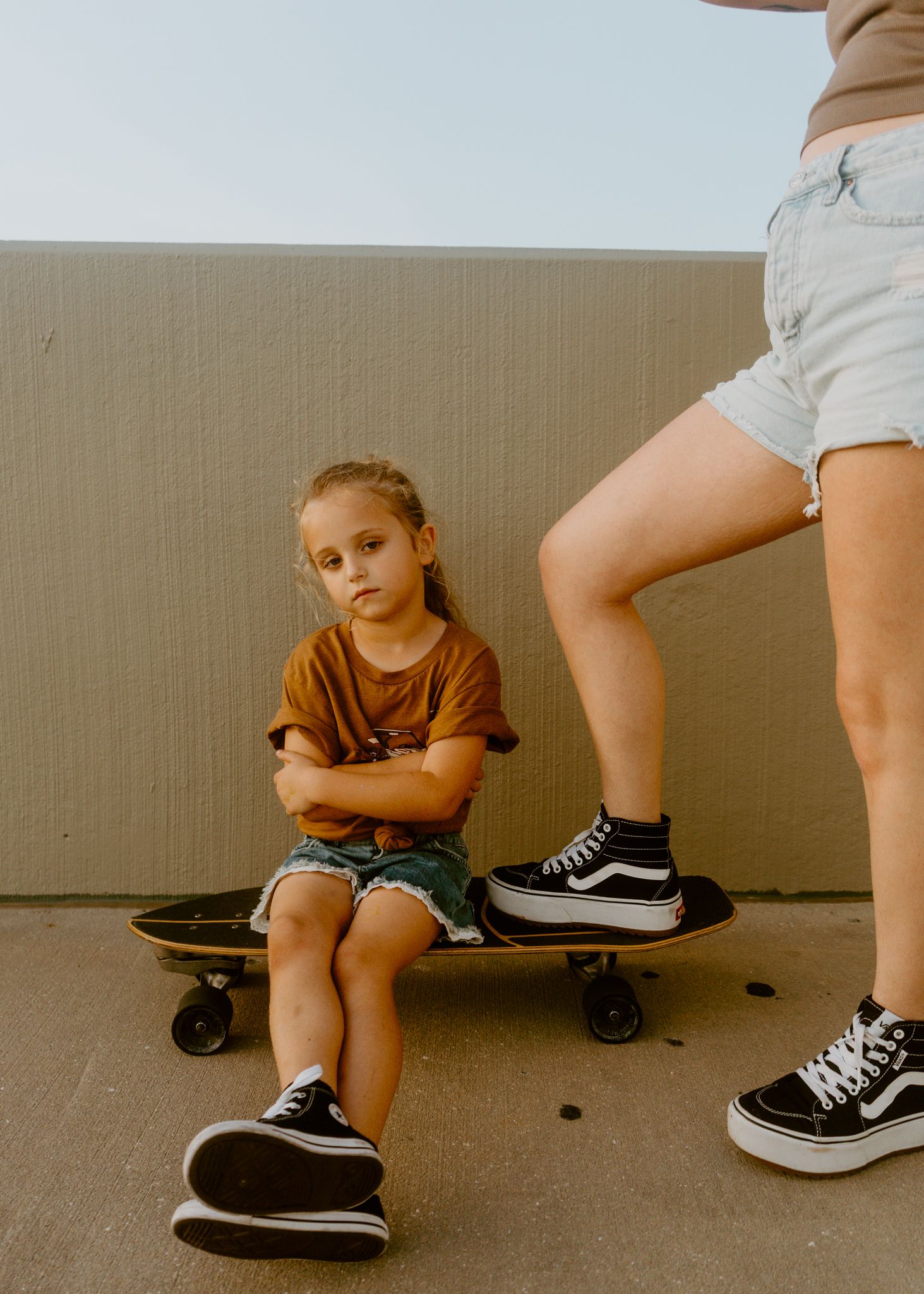 Young girl sitting cross-armed on a skateboard beside another person wearing Vans sneakers outdoors.