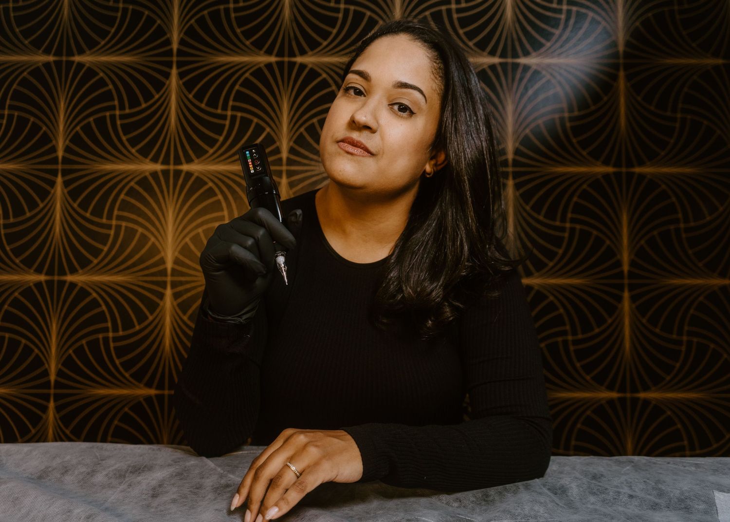 A tattoo artist in black gloves holds a tattoo machine, seated at a marble table against a gold patterned wall.
