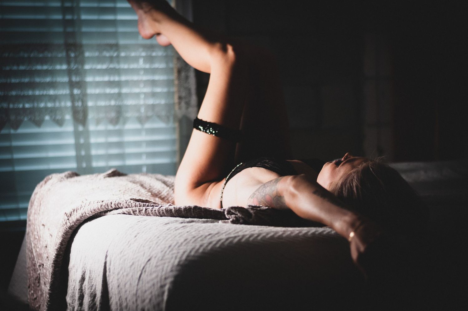 A woman in lingerie lies face down on a bed in a dimly lit room with soft light filtering through blinds.