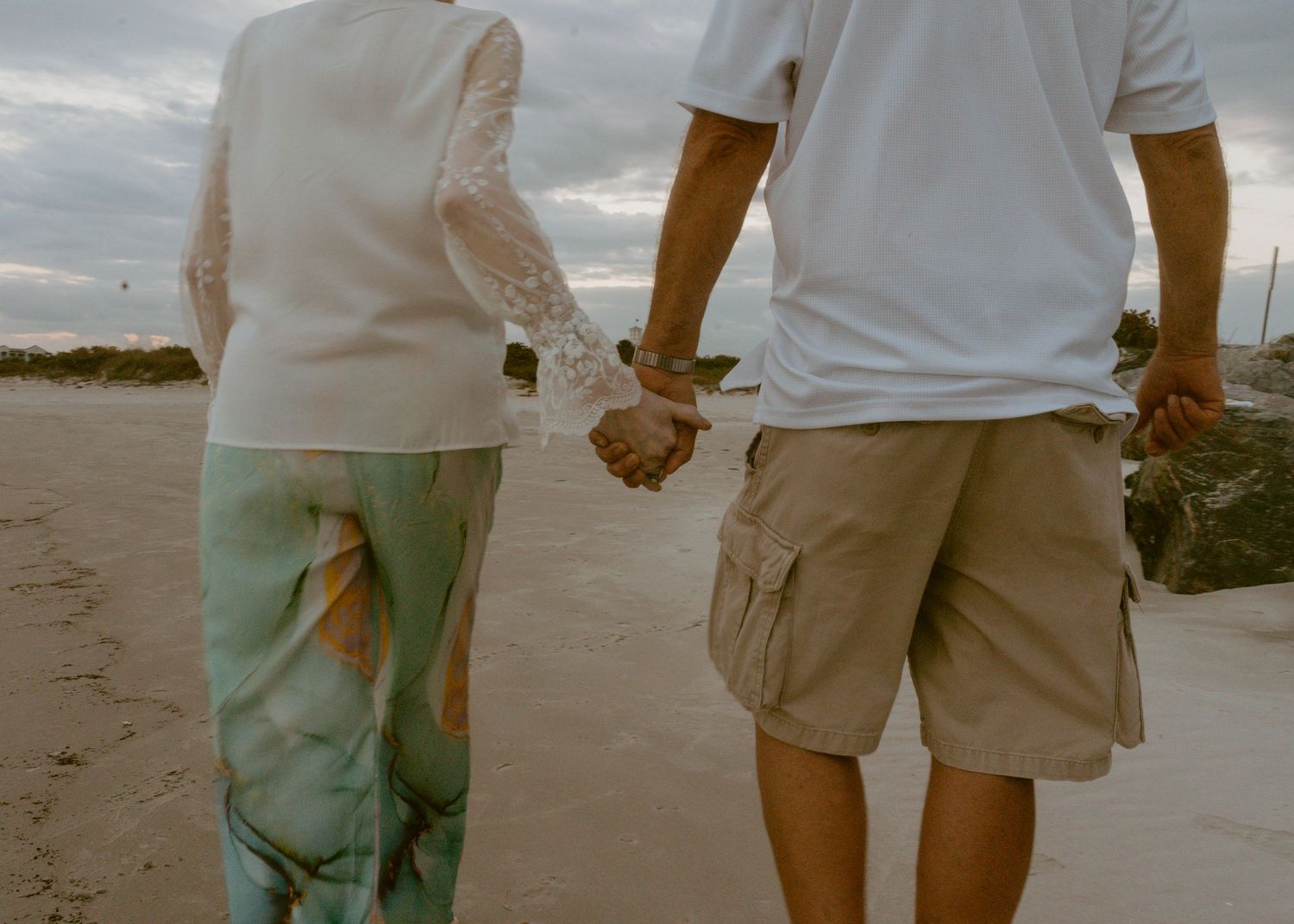 Elderly couple holding hands while walking on a sandy beach path, wearing casual summer clothing on a cloudy day.