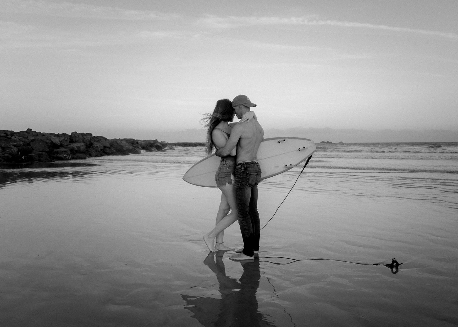 Romantic couple kissing on beach at sunset holding surfboards in black and white photography.